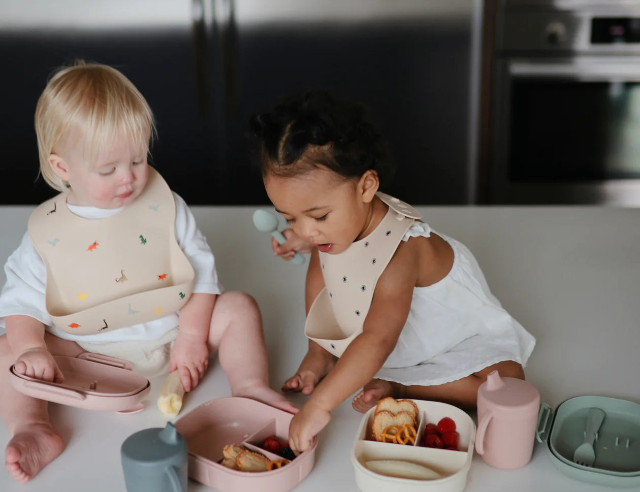 Two toddlers eating at a table wearing Mushie Silicone Baby Bibs