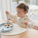 Baby eating at a table while wearing the Mushie Silicone Baby Bib in Whales