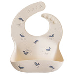 Mushie Silicone Baby Bib in Whales, a cream bib with a blue whale print and a catch pocket