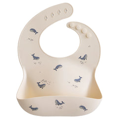 Mushie Silicone Baby Bib in Whales, a cream bib with a blue whale print and a catch pocket