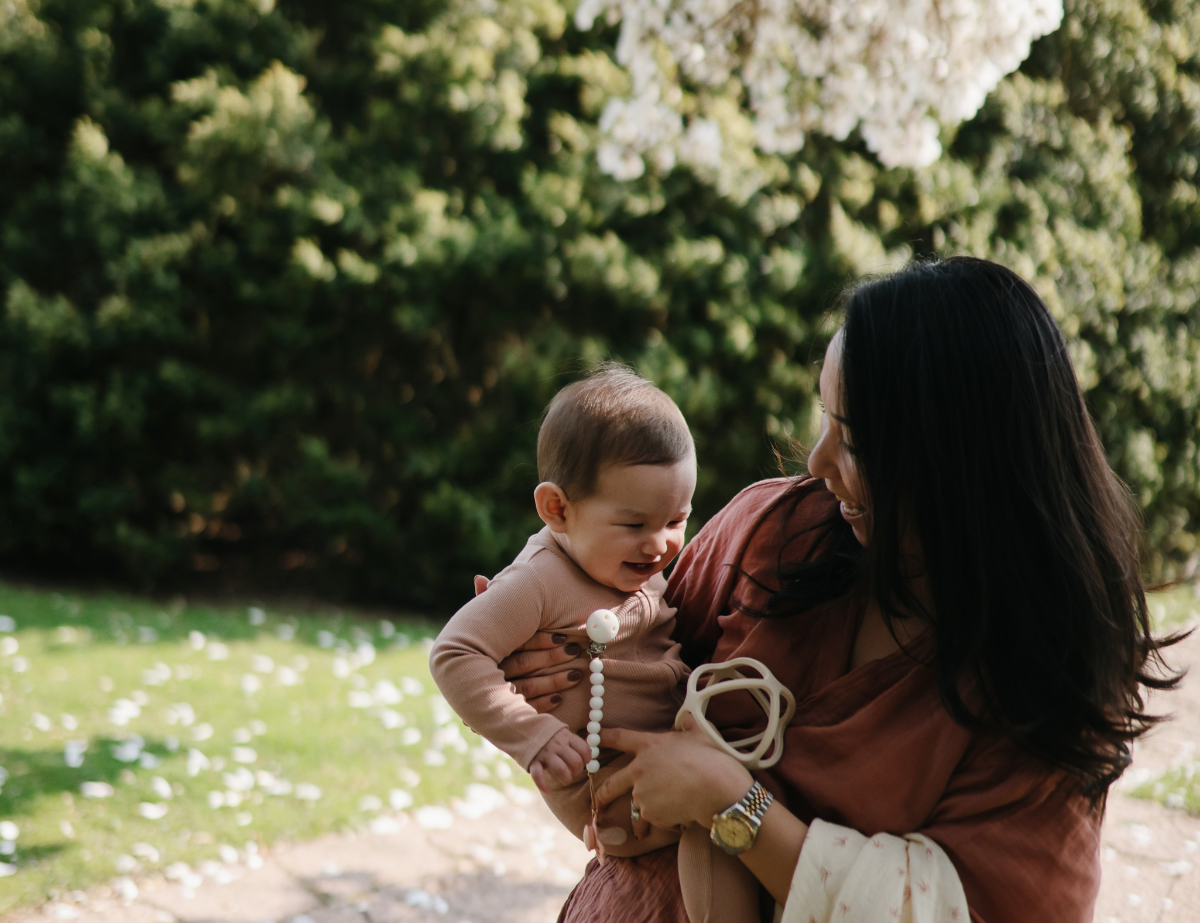 Mother holding a baby under a blossoming tree with a Mushie Silicone Pacifier Clip
