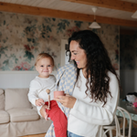 Mother holding a toddler with a Mushie Silicone Pacifier Clip in a floral living room