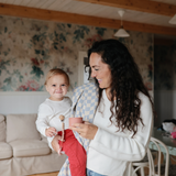 Mother holding a toddler with a Mushie Silicone Pacifier Clip in a floral living room