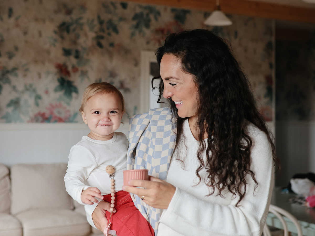 Mother holding a toddler with a Mushie Silicone Pacifier Clip in a floral living room