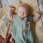 Sleeping baby in a bonnet with a Mushie Silicone Pacifier Clip on a green muslin blanket