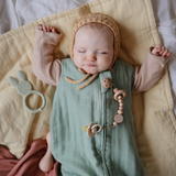Sleeping baby in a bonnet with a Mushie Silicone Pacifier Clip on a green muslin blanket