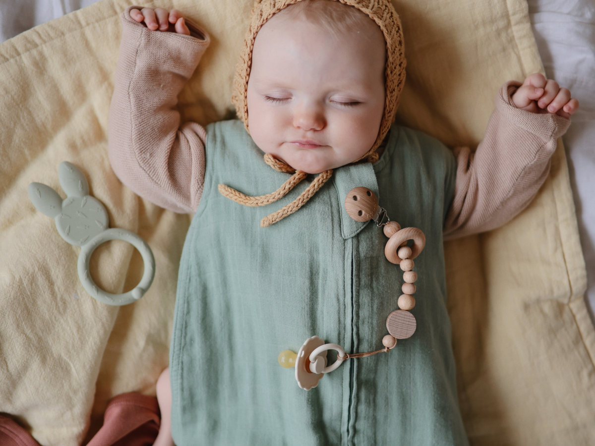 Sleeping baby in a bonnet with a Mushie Silicone Pacifier Clip on a green muslin blanket
