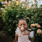 Smiling toddler in a pink dress with a Mushie Silicone Pacifier Clip in a flower garden