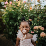 Smiling toddler in a pink dress with a Mushie Silicone Pacifier Clip in a flower garden