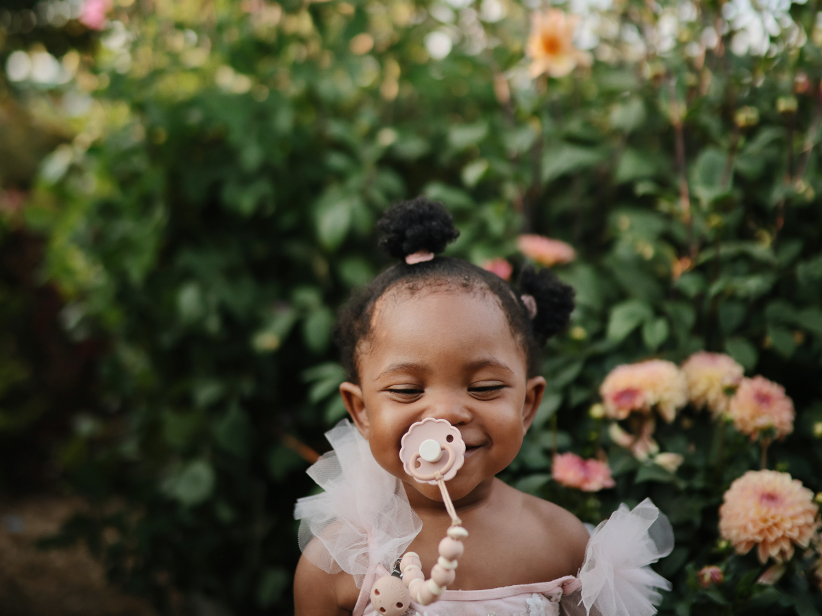 Smiling toddler in a pink dress with a Mushie Silicone Pacifier Clip in a flower garden