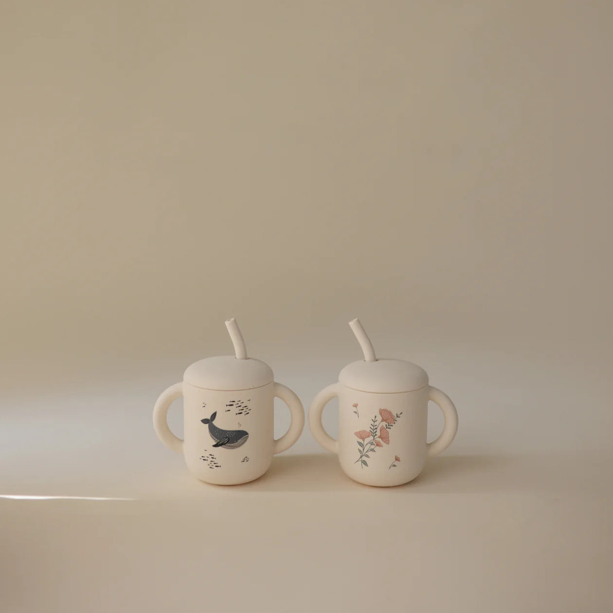 Mushie Silicone Training Cups in Whale and Pink Flowers side by side on a neutral surface
