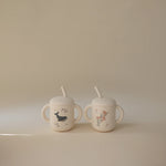 Mushie Silicone Training Cups in Whale and Pink Flowers side by side on a neutral surface