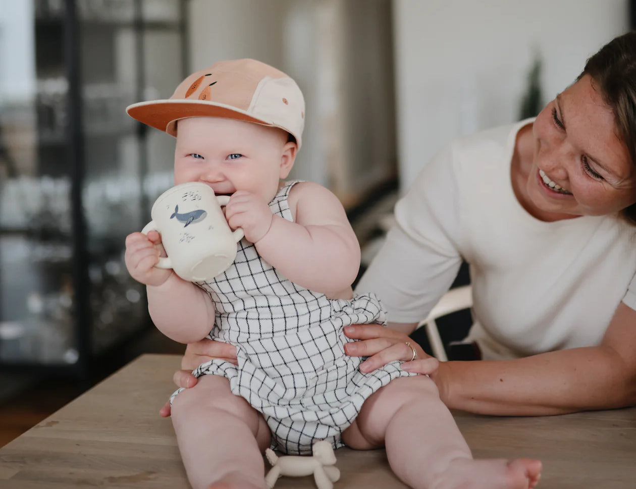 Baby in a cap drinking from the Mushie Silicone Training Cup in Whale while a parent smiles