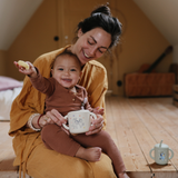 Mother holding a smiling baby with a Mushie Snack Cup in Bluey Ivory in a sunlit room