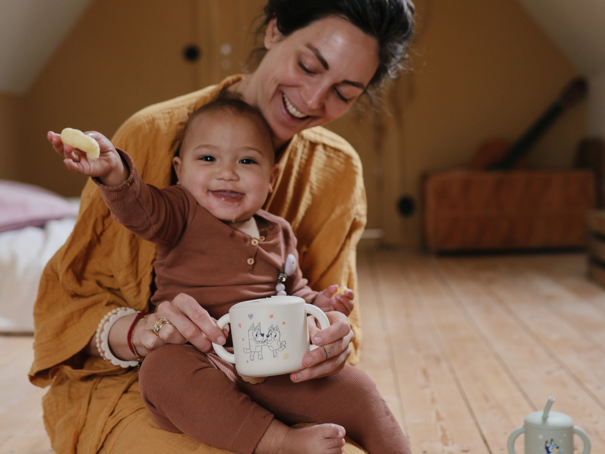Mother holding a smiling baby with a Mushie Snack Cup in Bluey Ivory in a sunlit room