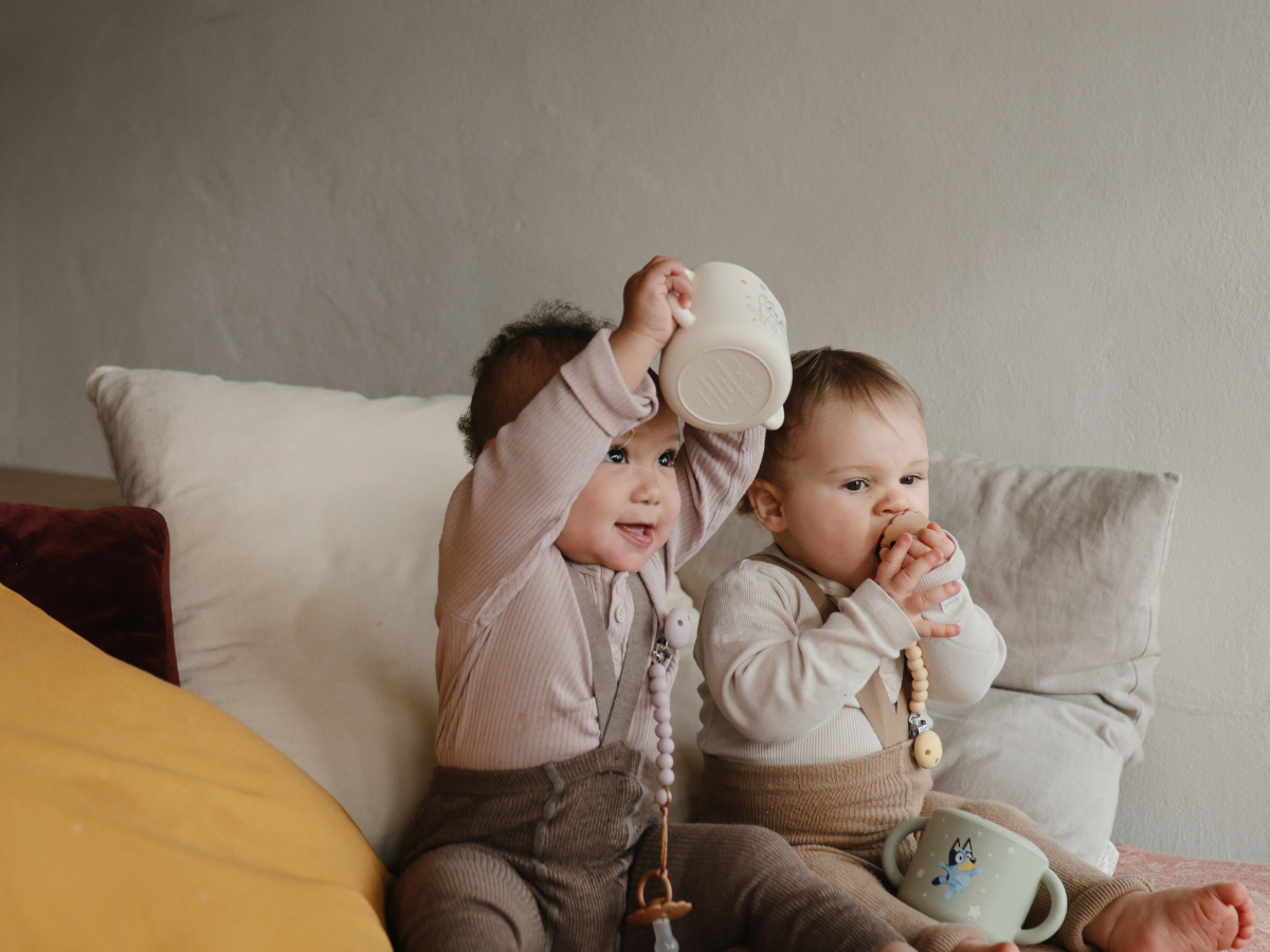 Toddler lifting a Mushie Snack Cup in Bluey Ivory overhead while sitting on a bed