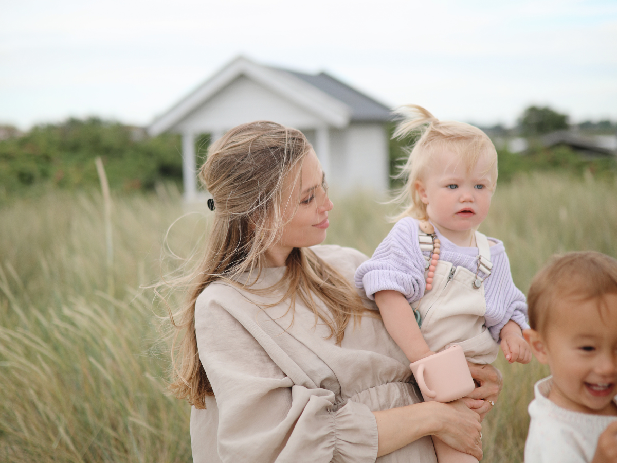 Mother holding a toddler with a Mushie Snack Cup in Blush in a grassy field