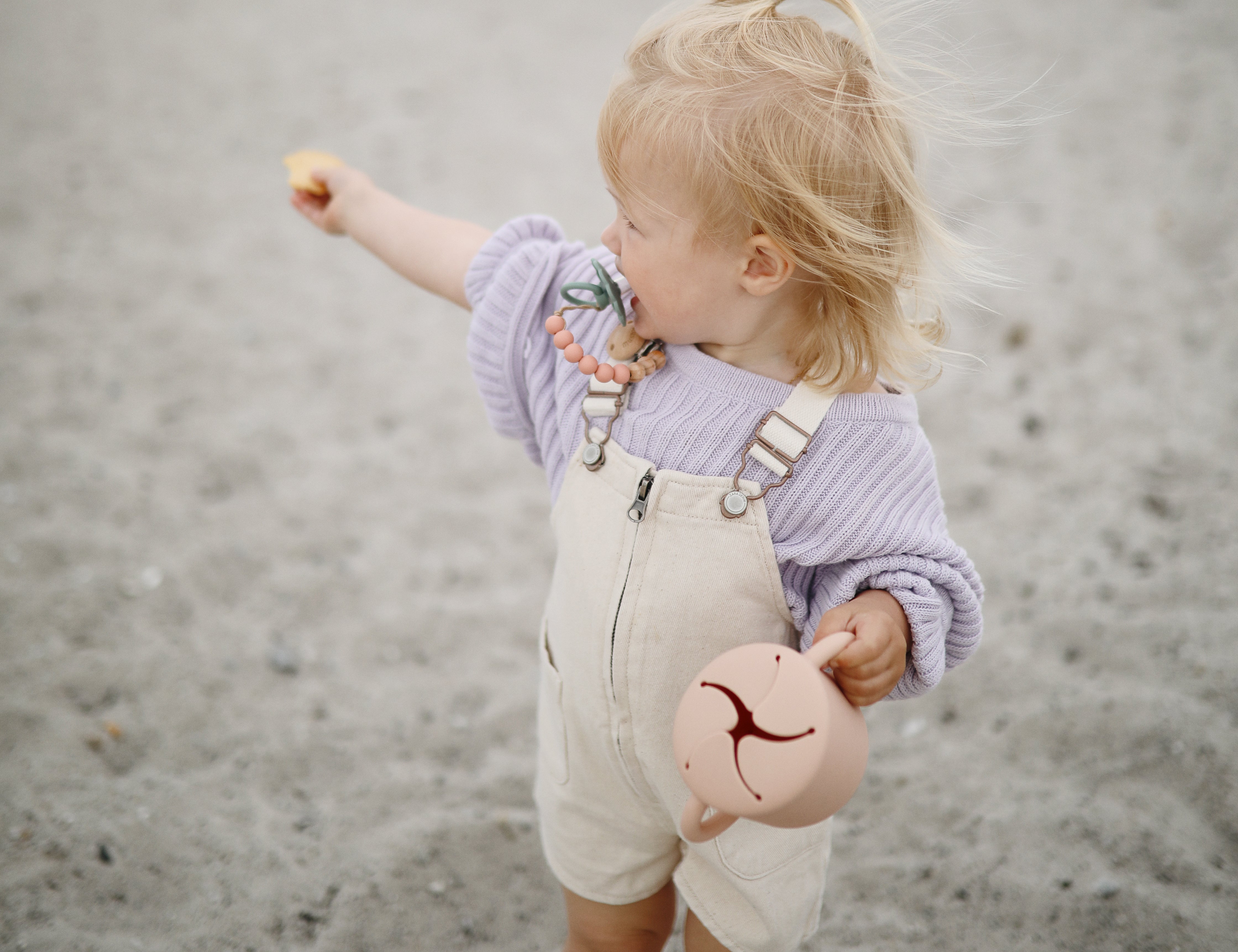 Toddler on a beach holding a Mushie Snack Cup in Blush