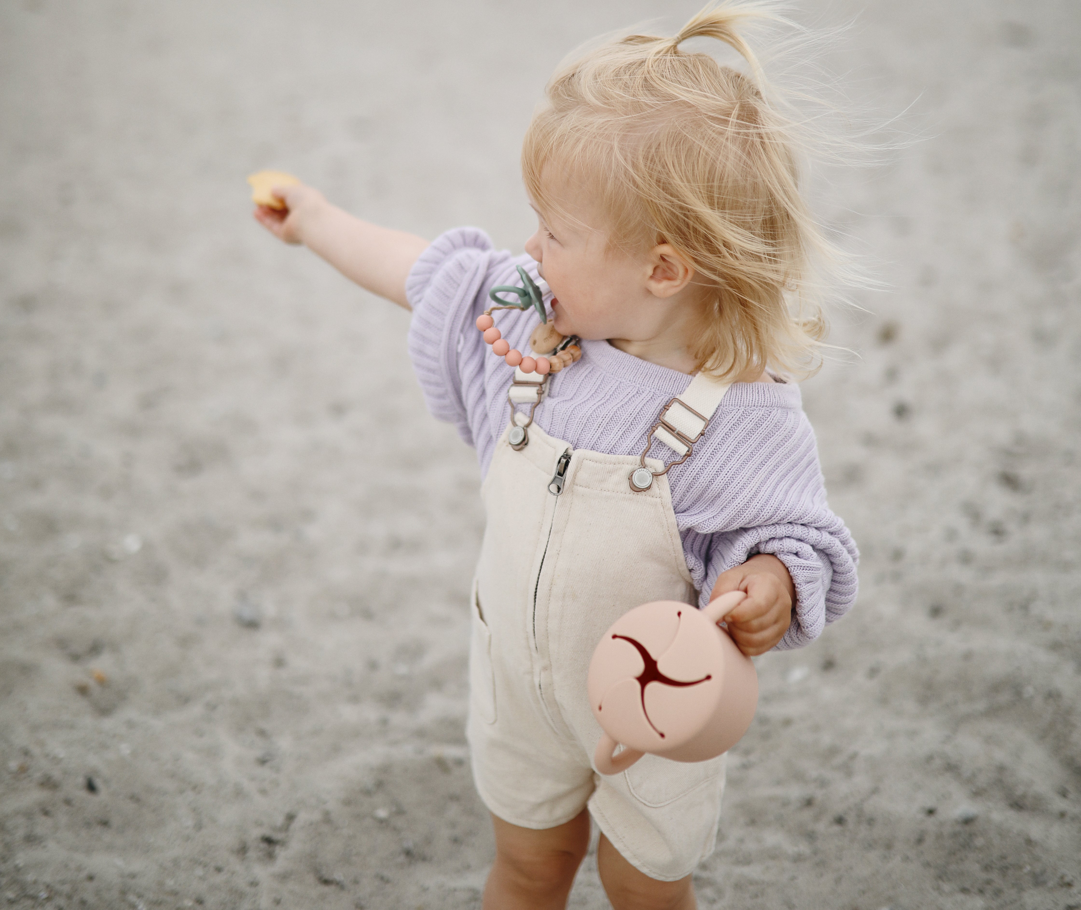 Toddler on a beach holding a Mushie Snack Cup in Blush