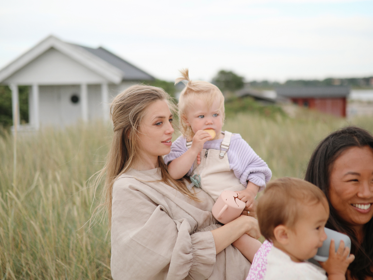 Two mothers with toddlers holding Mushie Snack Cups on a grassy beach