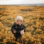 Toddler in a bonnet and plaid coat holding a Mushie Snack Cup in a field of yellow flowers