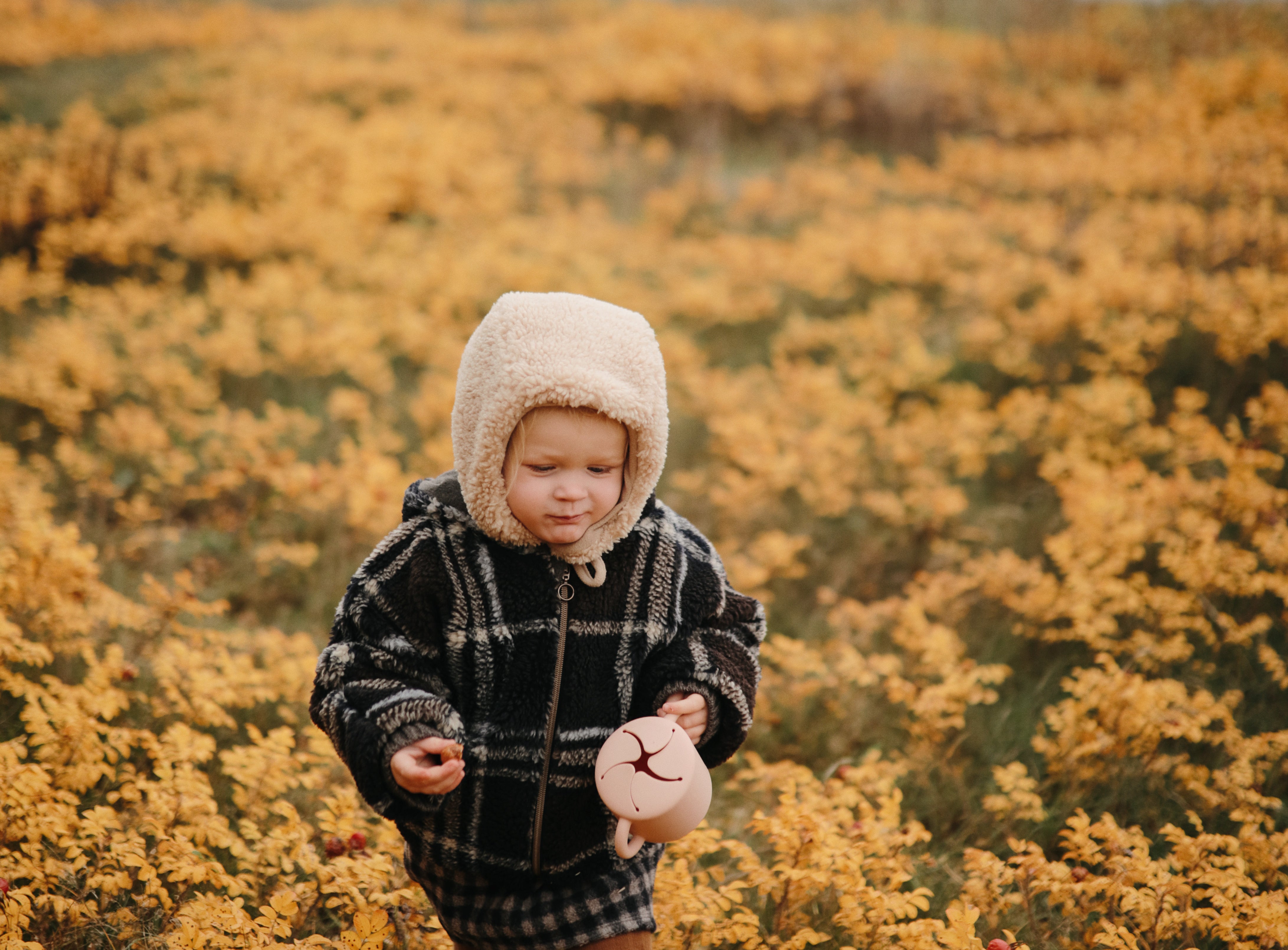 Toddler in a bonnet and plaid coat holding a Mushie Snack Cup in a field of yellow flowers
