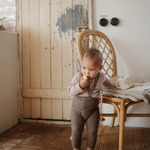 Toddler eating a snack while holding a Mushie Snack Cup beside a rattan chair