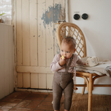 Toddler eating a snack while holding a Mushie Snack Cup beside a rattan chair