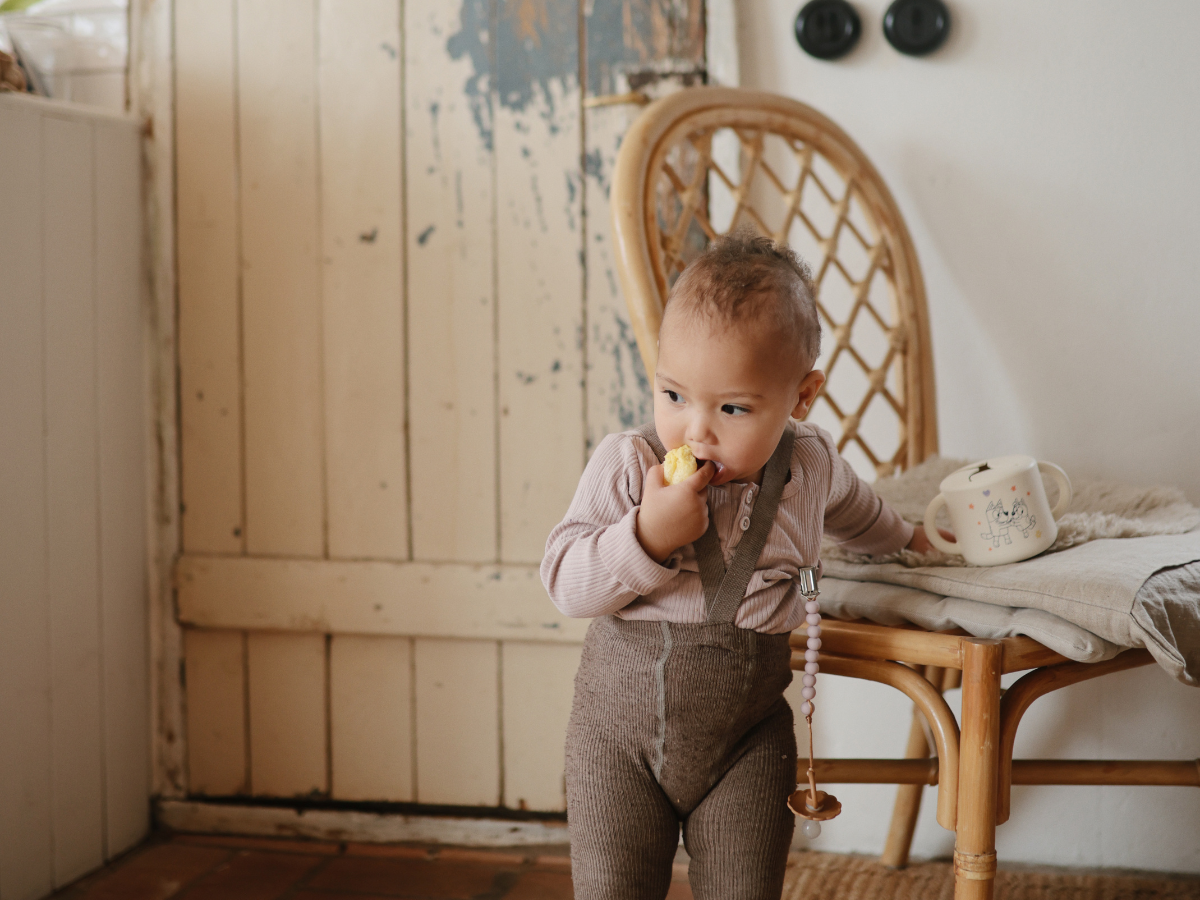 Toddler eating a snack while holding a Mushie Snack Cup beside a rattan chair