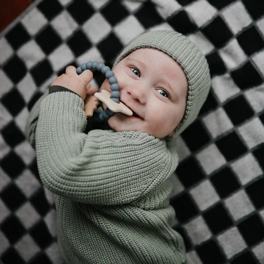 Baby in a green knit outfit chewing the Mushie Space Teething Ring on a checkered blanket