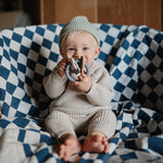 Baby in a cream knit outfit holding the Mushie Space Teething Ring on a blue checkered blanket