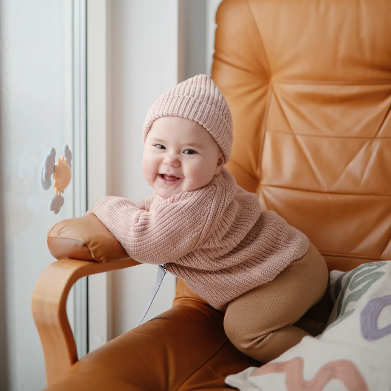 Smiling baby in a pink knit set on a leather chair with the Mushie Sunshine Suction Spinner Toy