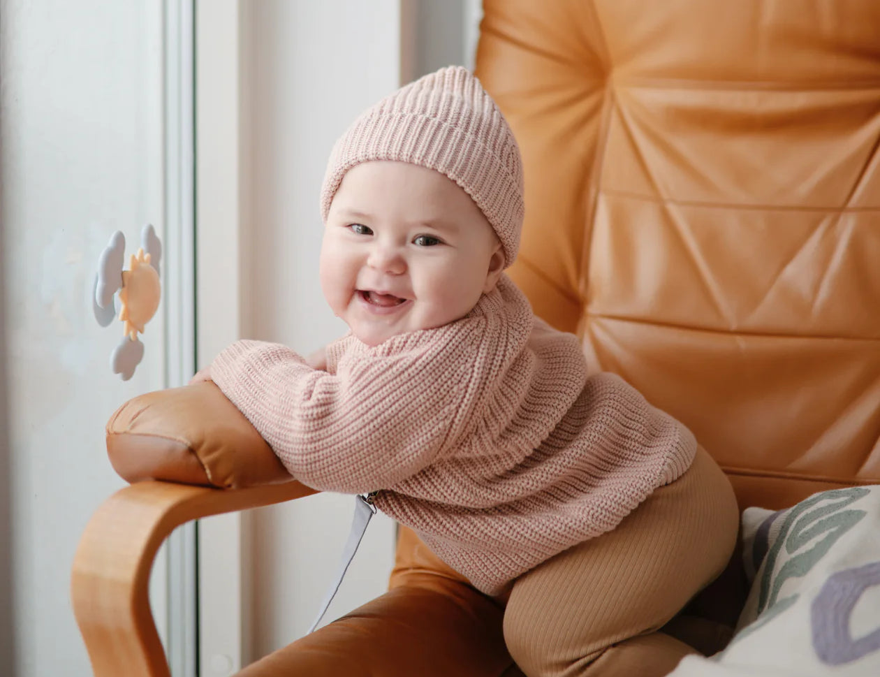 Smiling baby in a pink knit set on a leather chair with the Mushie Sunshine Suction Spinner Toy