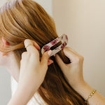 A woman slides the Mulberry Floral oval lay flat claw clip into her long hair in soft window light.