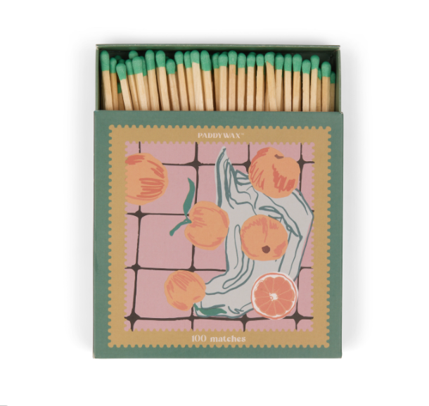 Green and pink Paddywax matchbox with oranges and a hand on a checkered ground, green match tips