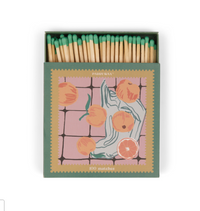 Green and pink Paddywax matchbox with oranges and a hand on a checkered ground, green match tips