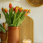 Paddywax Tulip Glass Candle lit on a shelf beside a vase of orange tulips