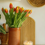 Paddywax Tulip Glass Candle lit on a shelf beside a vase of orange tulips