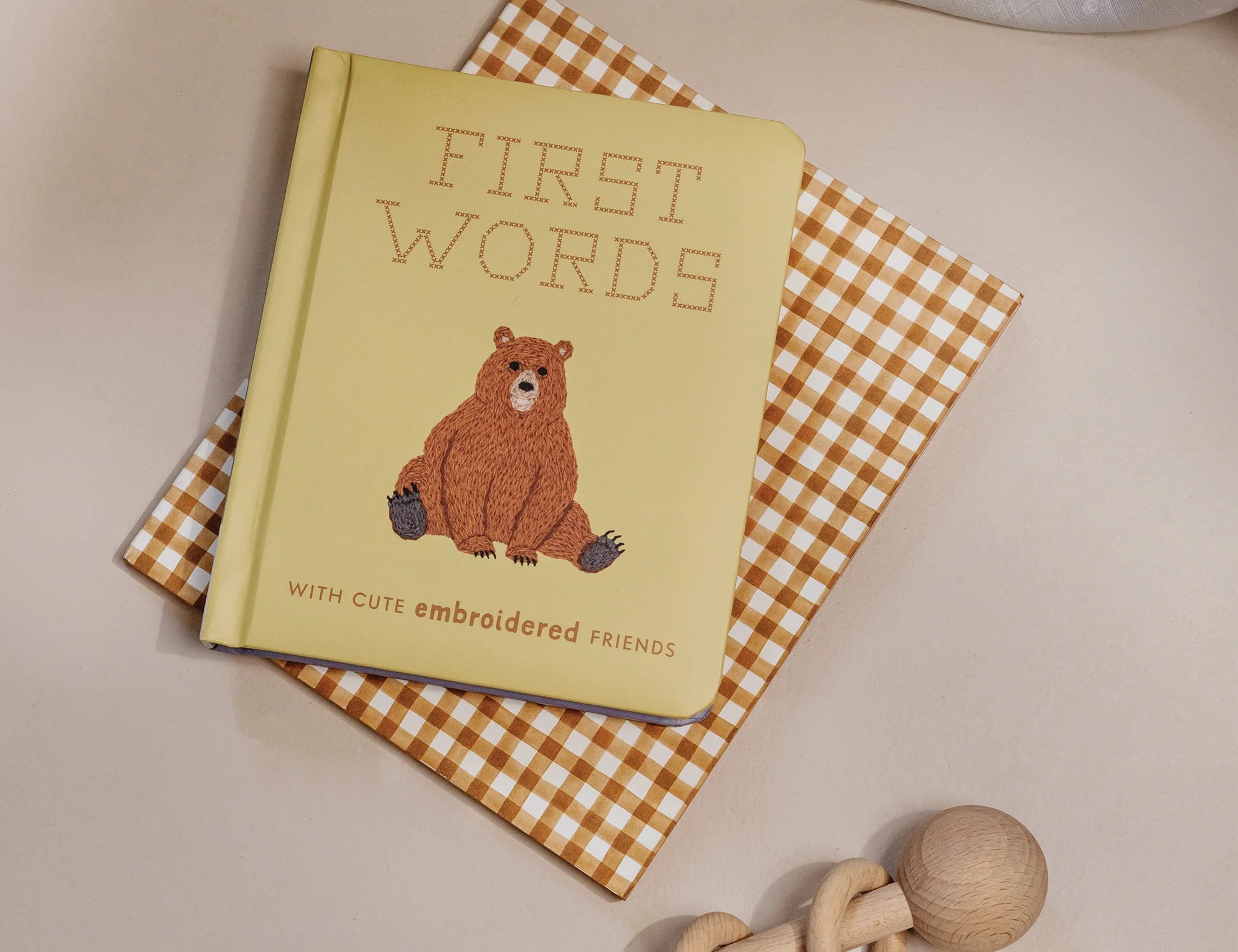 Paige Tate & Co First Words Embroidered Friends Book on a gingham cloth with a wooden rattle