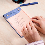 Hands writing a list on the Papier Fabric Hardcover Notepad in pink with a blue pen