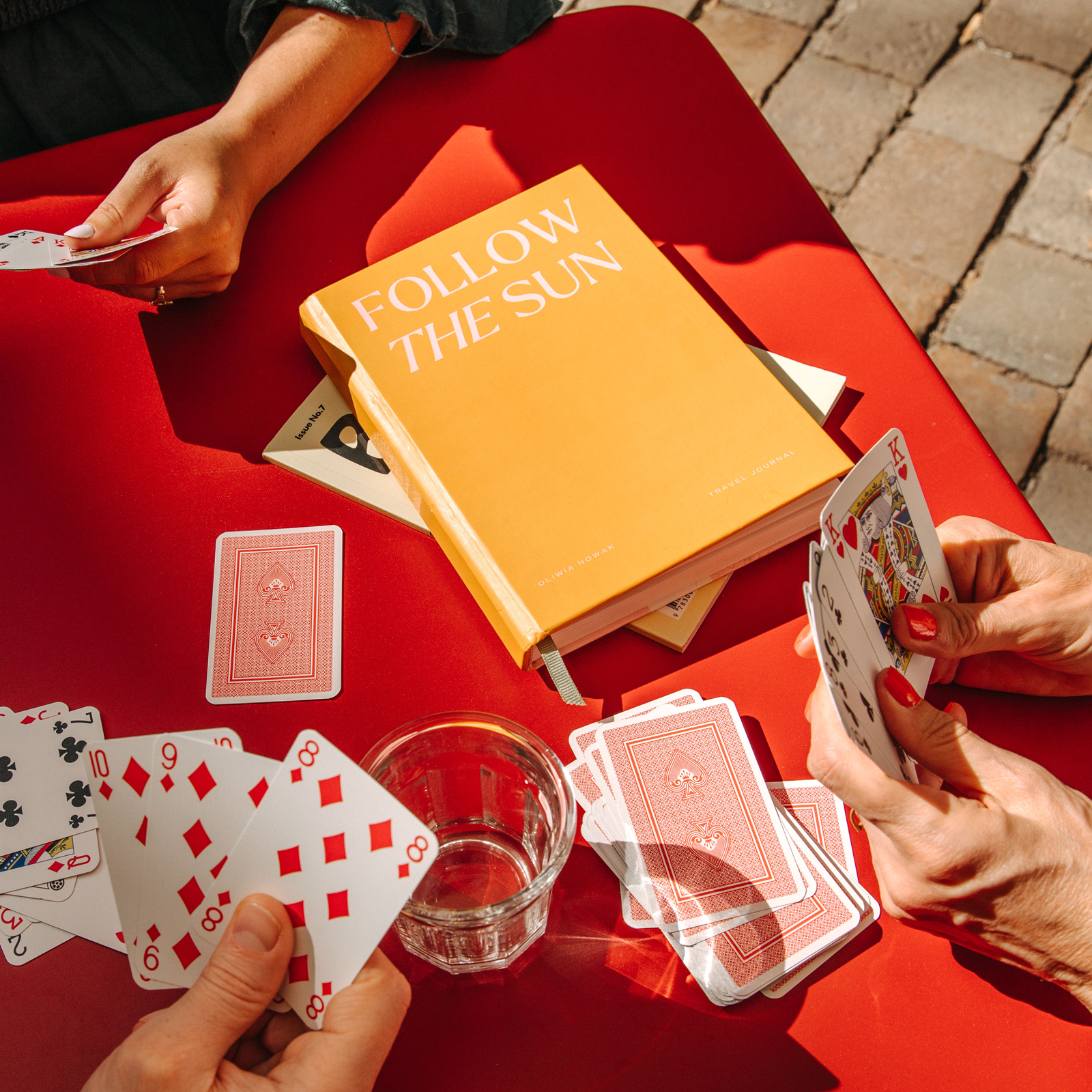 Papier Follow the Sun Travel Journal on a red table with playing cards and a drink