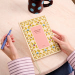 Hands holding the Papier Guided Gratitude Journal A5 Folk Garden at a table with a pen