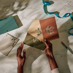 Hands writing on Papier Keep in Touch Notecards with a teal ribbon and rust envelopes