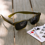 Peepers 18th Hole Sunglasses in olive green resting on a wooden table next to playing cards