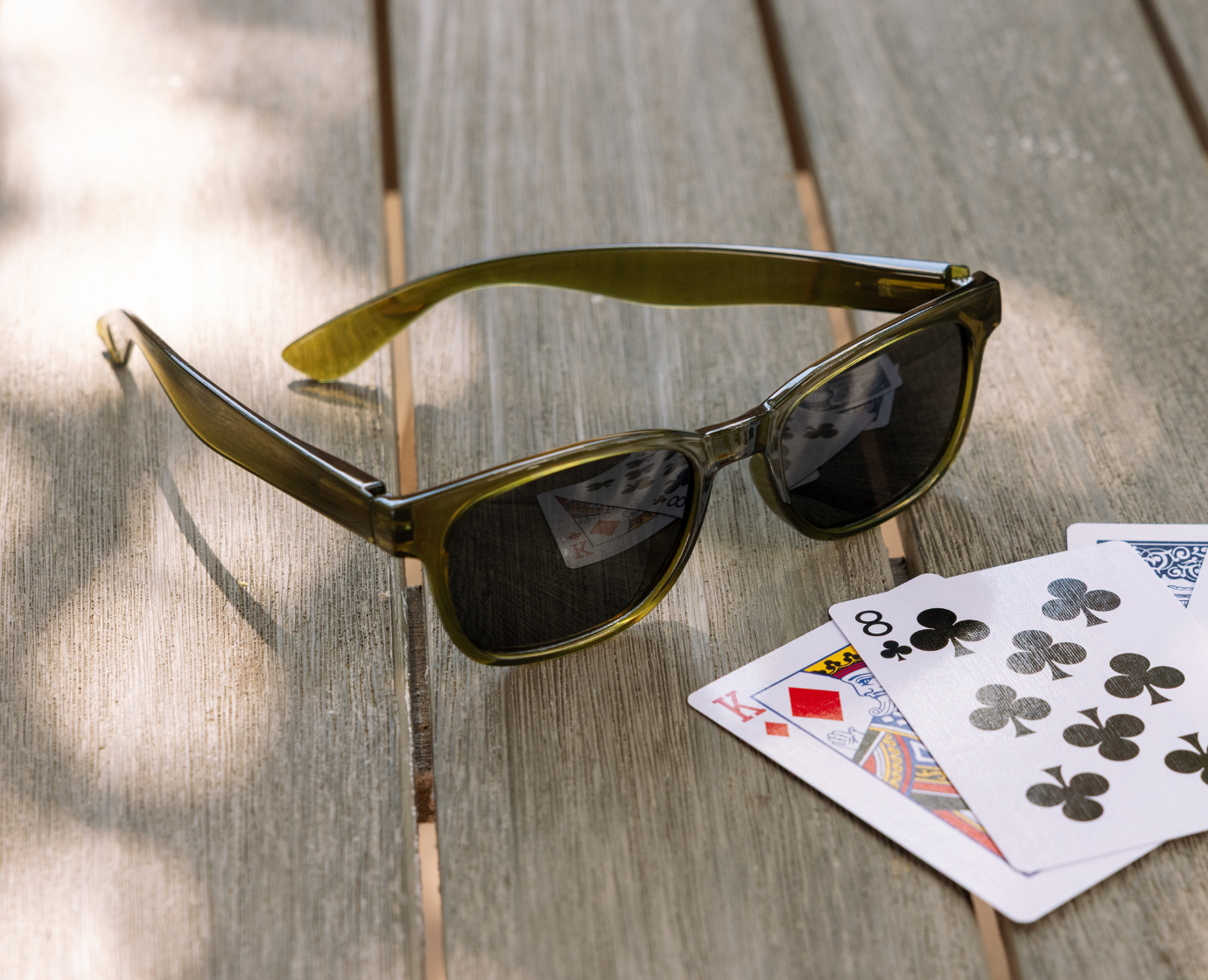 Peepers 18th Hole Sunglasses in olive green resting on a wooden table next to playing cards