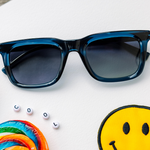 Peepers Airplane Mode Sunglasses in navy on a white surface with a rainbow lollipop, smiley patch and letter beads spelling COOL