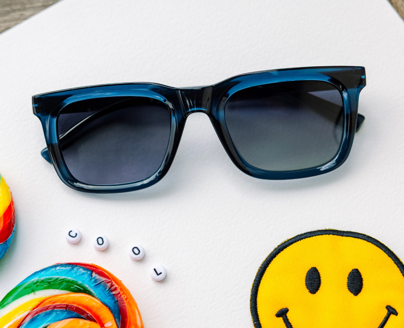 Peepers Airplane Mode Sunglasses in navy on a white surface with a rainbow lollipop, smiley patch and letter beads spelling COOL