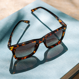 Peepers Airplane Mode Sunglasses in tortoise resting on a blue linen cloth in sunlight