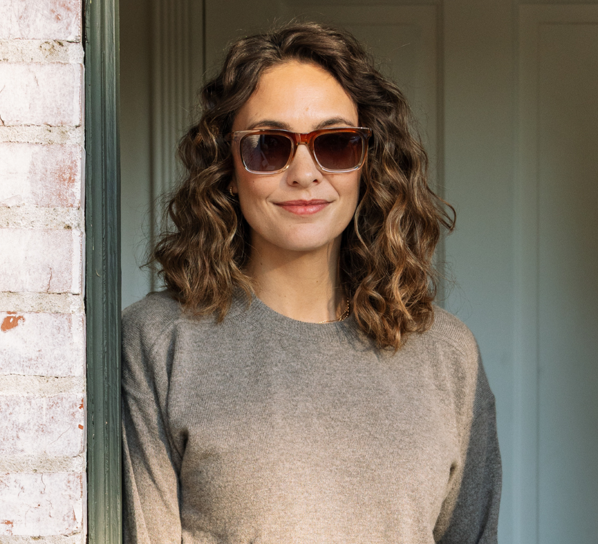Peepers Airplane Mode Sunglasses in tortoise worn by a curly-haired woman in a taupe sweater in a doorway