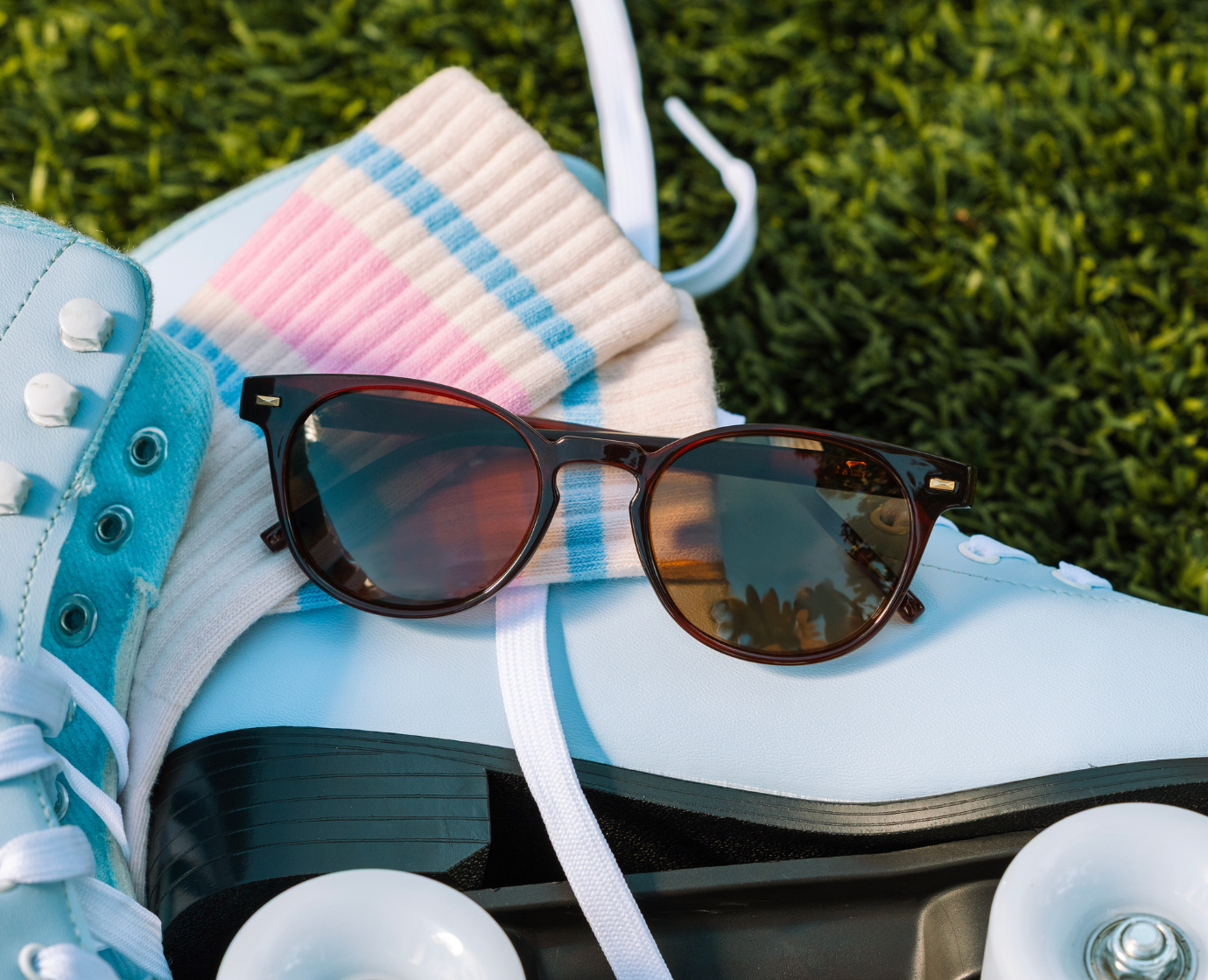 Peepers Boho II Sunglasses in Brown resting on a pair of light blue roller skates in the grass
